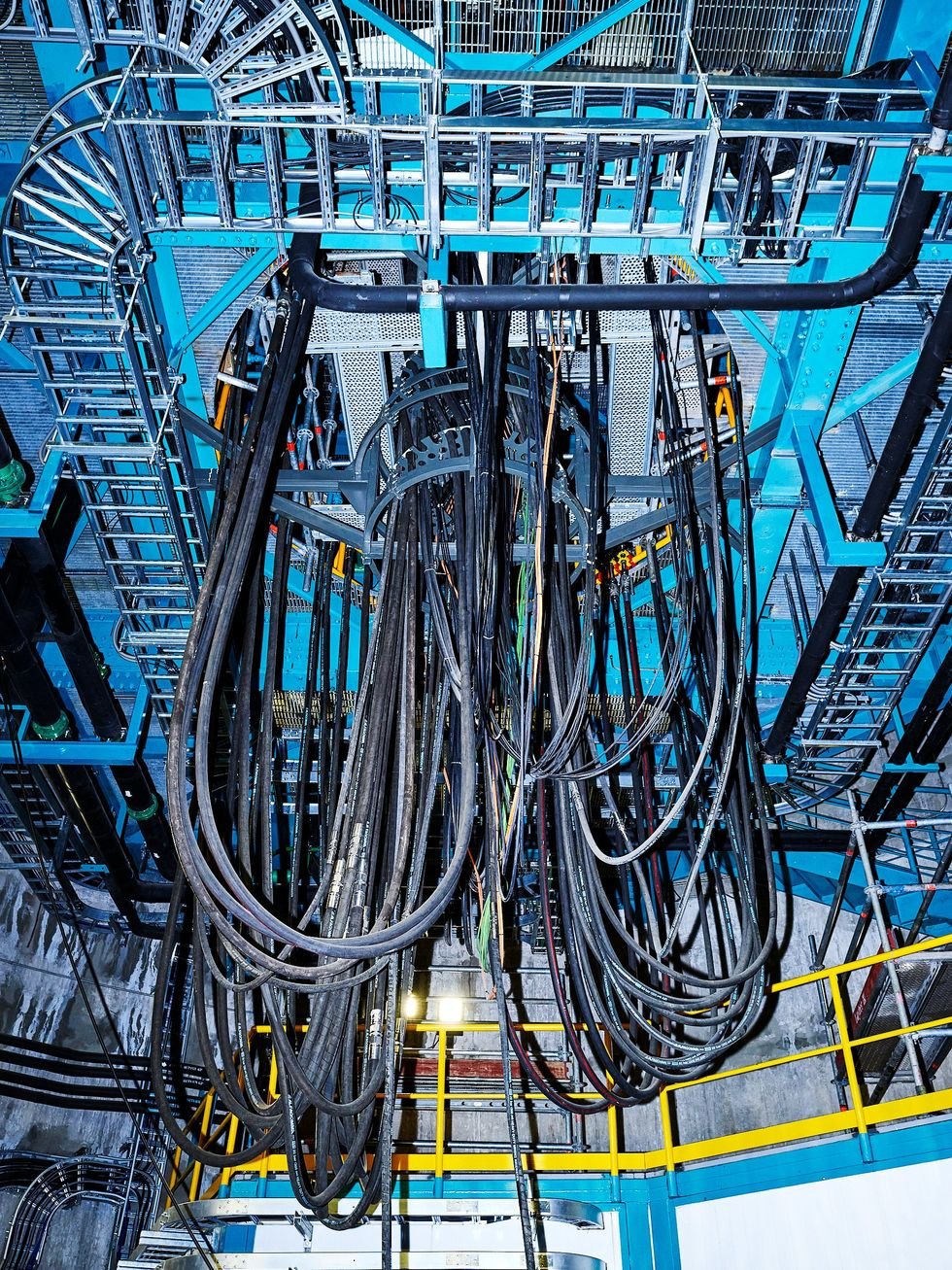 Numerous thick cables hang in an industrial setting, surrounded by blue metal scaffolding.