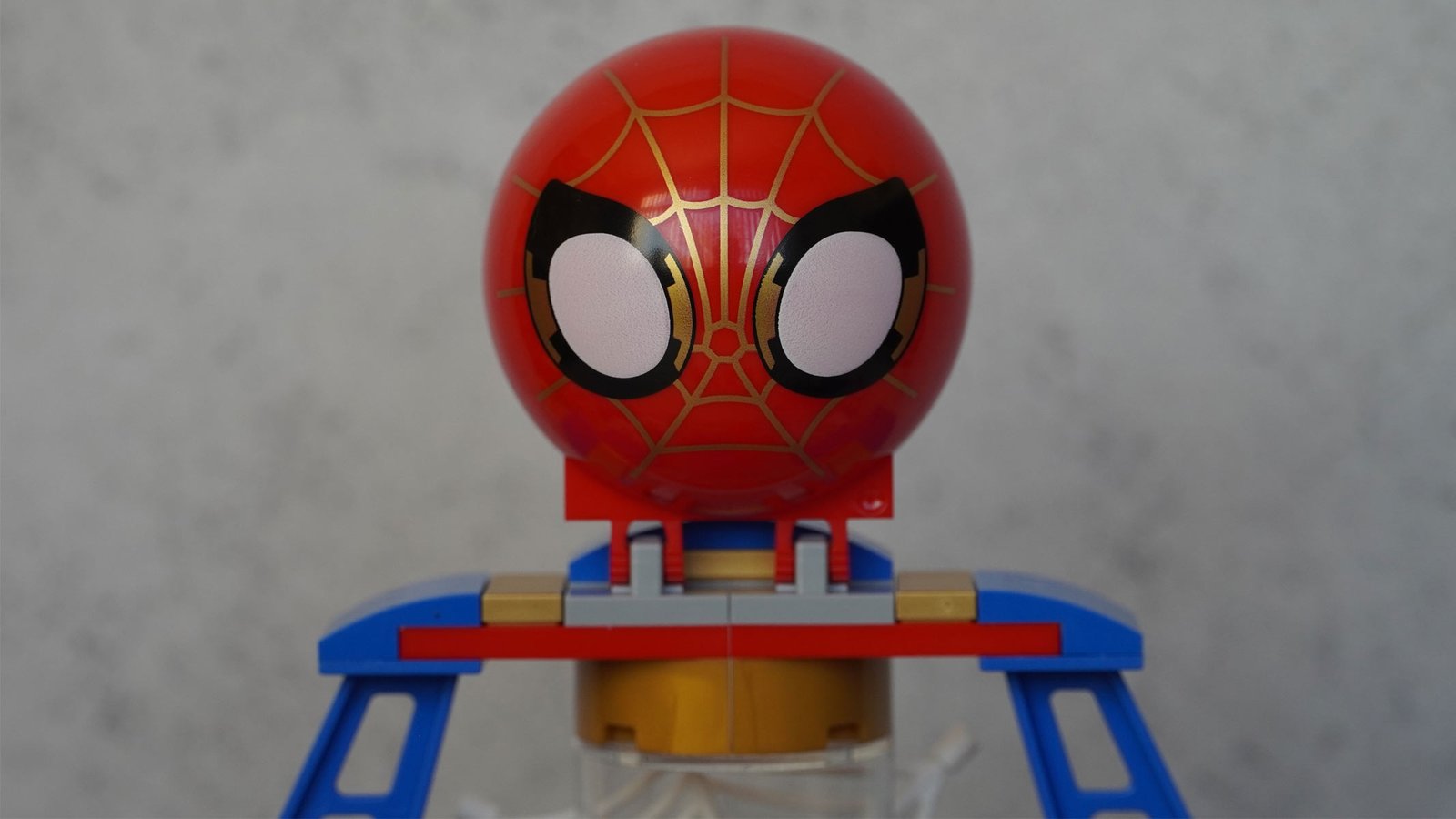 A large round print of Spider-Man's face features large white eyes with black around the side on a red and gold face, all on a grey background.