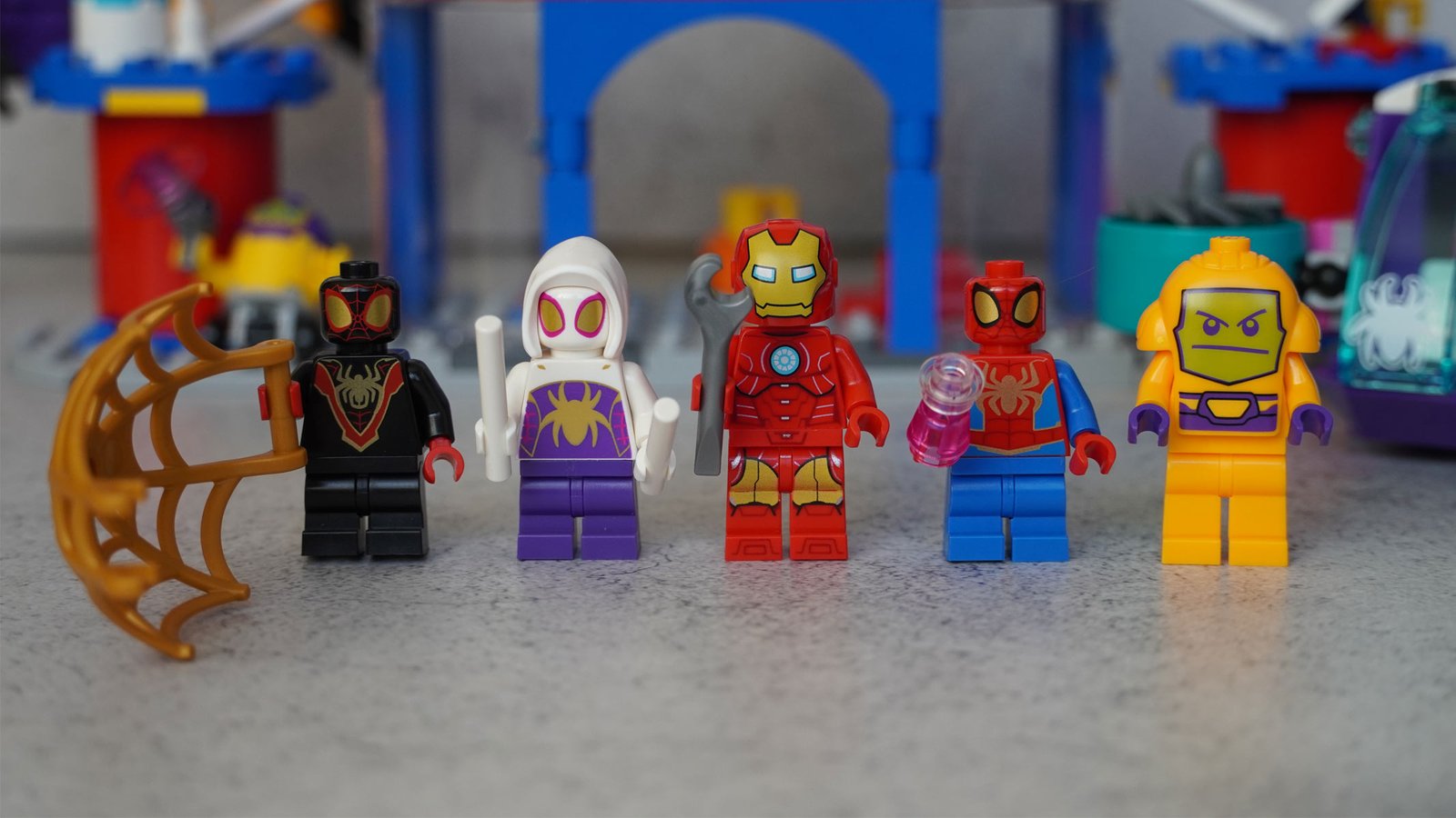 Five minifigures, one of Spider-Man in black, one in purple with a white hood and Spider-Man in red and blue colors. Iron Man and a yellow figure with an alien face also feature on a grey background.