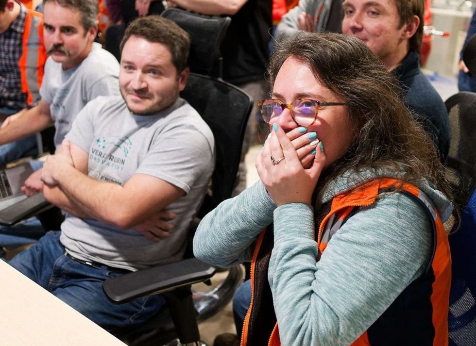 Group of people seated in office chairs look at a screen (not shown) and smile slightly, with one person covering their mouth with their hands.