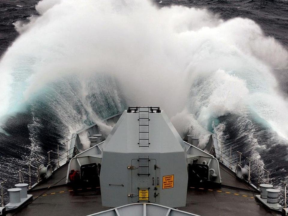 View from a naval ship’s deck as it cuts through large waves during high-storm conditions.